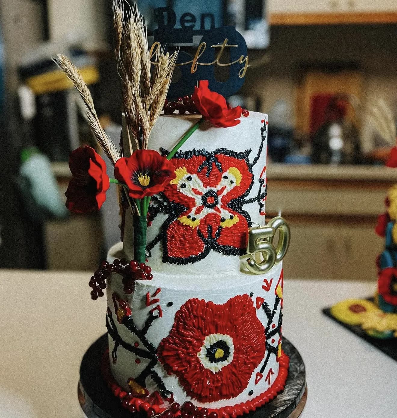 Two-tier white cake with hand-piped red poppies and black branches plus dried wheat and gold 50