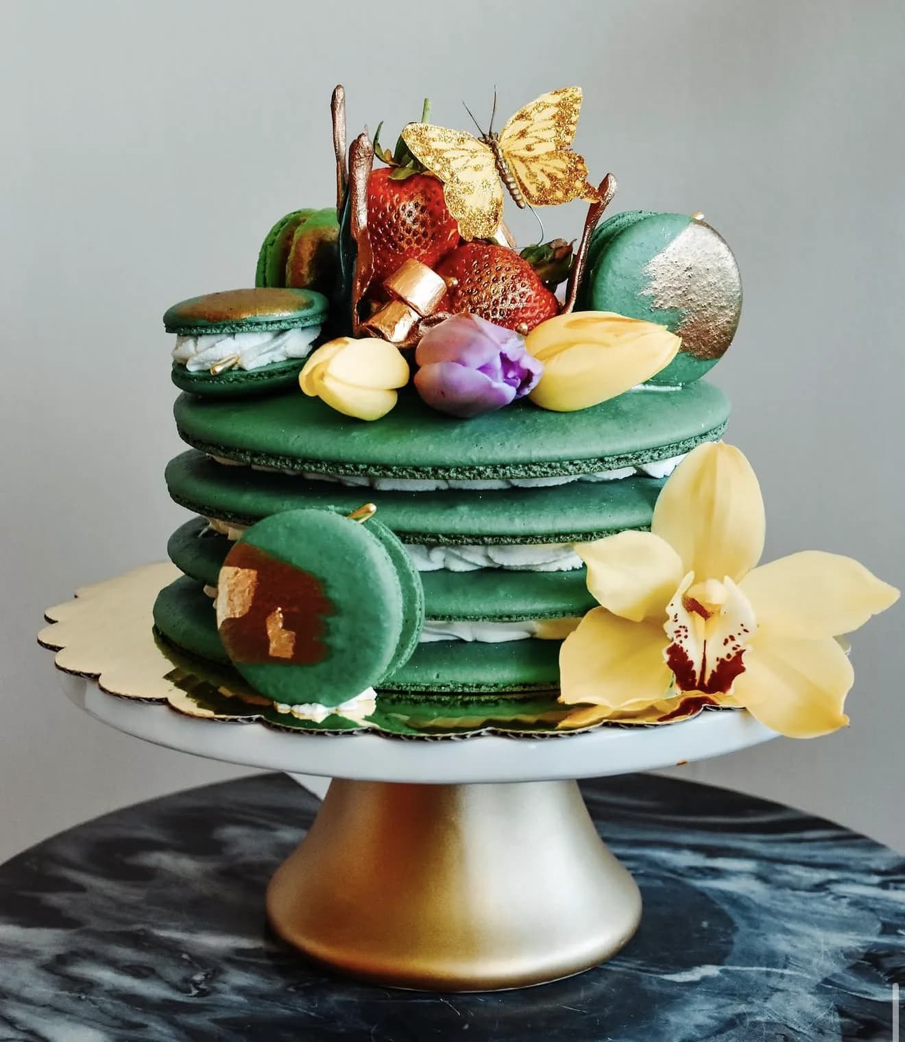 Stacked emerald green mega macaron cake topped with strawberries gold butterfly and orchid
