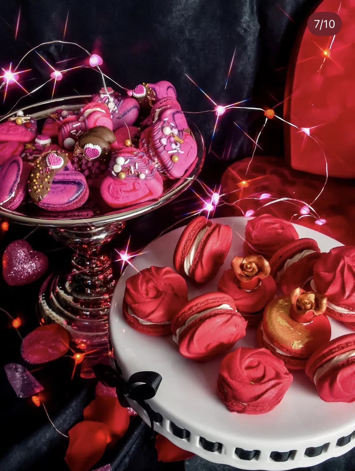 Red rose-shaped macarons with gold sugar sphere accents on white cake stand with fairy lights
